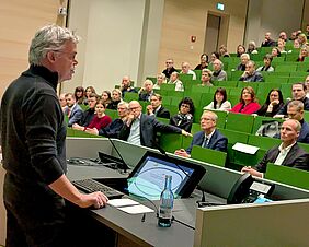 Ein Mann steht an einem Rednerpult in einem Hörsaal. Die Kamera befindet sich direkt hinter ihm, wodurch ein Blick auf die grünen Stühle im Hörsaal frei wird. Viele der Plätze sind durch Personen besetzt. Das Publikum hört gespannt zu.