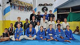 Auf diesem Gruppenfoto sind Trainer, Kinder und Jugendliche eines Jiu-Jitsu-Kurses zu sehen. Die Personen haben sich in drei Reihen positioniert und tragen klassische Kampfsportroben in schwarz, weiß und blau. Das Foto wurde wahrscheinlich in einer Sport- oder Trainingsstätte aufgenommen. An den Wänden befinden sich Auszeichnungen wie Medaillen und Meistergürtel.