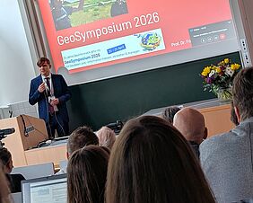 In einem Hörsaal spricht eine Person zu den Anwesenden. Der Moderator trägt einen förmlichen, dunkelblauen Anzug. Hinter ihm erscheint eine Präsentation mit dem Titel „GeoSymposium 2026“.