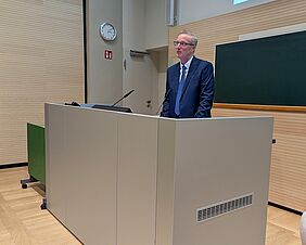 Ein Redner steht an einem Pult vor einer grünen Tafel. Er trägt einen blauen Anzug und eine blaue Krawatte.