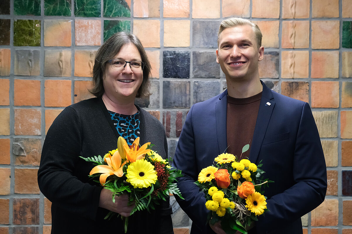 Auf dem Foto zu sehen sind Prof.in Dr.in Claudia Vogel, Vorsitzende des Erweiterten Senats und Constantin Friz, Studentischer Prorektor der Hochschule Neubrandenburg. Beide stehen im oberen Foyer von Haus 1, lächeln in die Kamera und halten jeweils einen Blumenstrauß in den Händen.