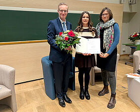 Drei Personen lächeln freundlich in die Kamera. Der Mann links außen hält einen üppigen Blumenstrauß in der Hand, die junge Frau in der Mitte des Bildes hält eine Urkunde.