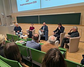 Fünf Personen sitzen während einer Podiumsdiskussion auf Stühlen in einem angedeuteten Halbkreis. Hinter ihnen befindet sich eine grüne Tafel, vor ihnen sitzt das Publikum auf Hörsaalbänken.